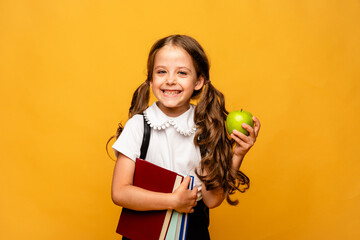 A cute little schoolgirl in a school uniform and with a school bag, books and green apple poses in the studio on a yellow background. Education. Place for text.