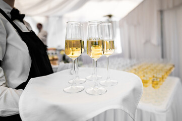 Close-up of a waiter’s hand holding a tray with four glasses of champagne, suggesting a celebration or event. More glasses are seen in the background