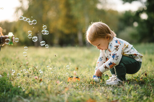 Boy catches soap bubbles in nature. Toddler little cute walking and playing in spring garden. 2 year old boy enjoying on green grass in park on sunny summer day. Outdoors creative activities for kids.
