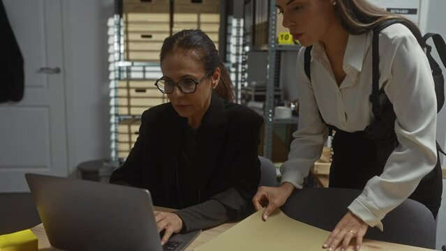 Two women in a well-lit office, with one sitting at a laptop while the other, in a holster, hands over a folder, implying they are detectives or investigators.