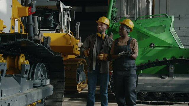 Caucasian male factory worker walking with African American female colleague along bulldozer and telling her about machine manufacturing process