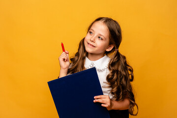 Clever little girl in school white shirt holding notebook and pen, smiling, thinking, doing homework, on a yellow background. back to school concept.