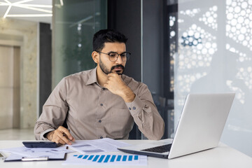 A serious young Indian man is sitting in the office at the desk, working with documents, looking...