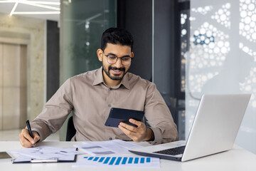 Smiling young Indian male accountant, financial expert and analyst working in office at laptop,...