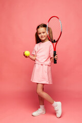 Cheerful little sports girl in pink sport suit playing tennis isolated over pink background. Sport, study, childhood concept. Copy space for ad, text. Beach summer tennis camp.