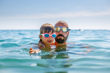 Father and daughter, waist-deep in a calm sea, wearing goggles, capturing a moment of beachside fun