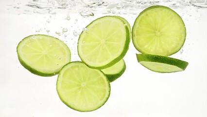 Slices fresh lime is thrown into water with splashes and bubbles filmed in slow motion on white background. Closeup view of green chopped ripe citrus floating in transparent aqua. Exotic fruit theme