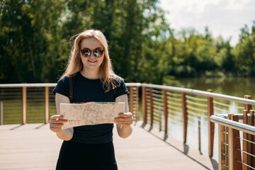 Stylish hipster woman in sunglasses holding paper map, wearing backpack and relaxing in nature. Travel and wanderlust concept. Amazing chill moment