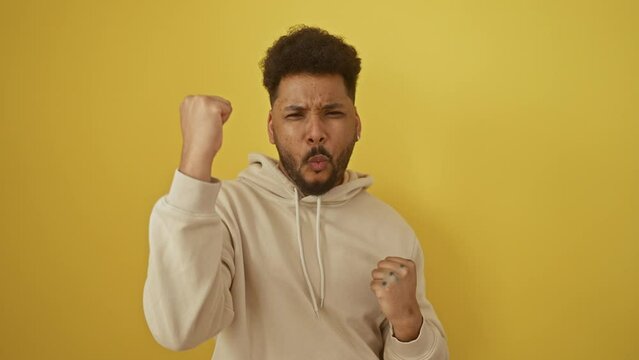 Joyful african american man, wearing hoodie, strikes a winning gesture! celebrating success, arms raised in triumph, surrounded by yellow isolated background! yes, a definite winner!