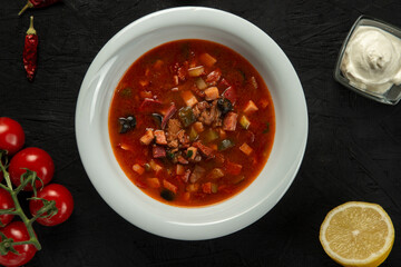 A bowl of delicious solyanka on a black background, soup with sausages and pickles. view from above