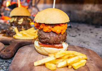 Delicious hamburger with bread, meat, cheddar cheese, bacon and sausage on top of wooden board, with french fries on the side. Hamburguer in front of fire.