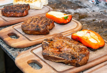 Several wooden boards with big pieces of grilled meat and red peppers with cheese on the side. 
