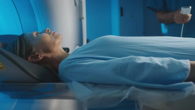 Slowmo close-up of middle-aged Caucasian woman in hospital gown lying on patients table moving into MRI machine under control of radiologist operating medical equipment by remote control