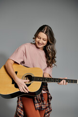 A woman in a pink t-shirt and a plaid shirt tied around her waist plays an acoustic guitar while smiling.