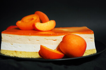 Cake made of cream and apricot jam next to apricots on a black plate on a dark background