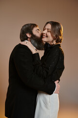 A couple dressed in sophisticated attire share a tender moment, the woman lovingly kissing the man on the cheek.