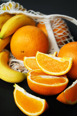 Whole and sliced ​​oranges next to bananas in a string bag on a dark background