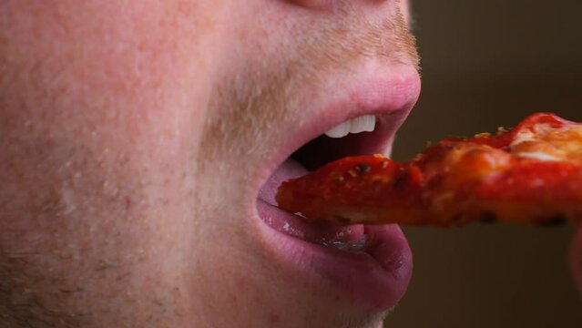 Close-up side view of a man's unshaven face, mouth bites off a piece of pepperoni pizza and chewing. Bright tasty delicious pizza. Concept of junk fast food. Unhealthy diet.