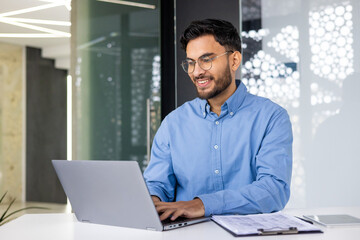 A smiling Indian man in a bib and blue shirt is working on a laptop, sitting at a desk in a modern...