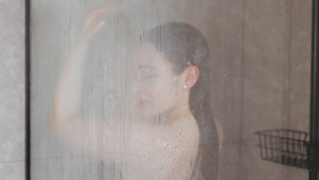 Showering behind frosted glass. A woman is seen taking a shower behind a frosted glass door The view is blurred due to the steam and water droplets on the glass, adding a sense of privacy and intimacy