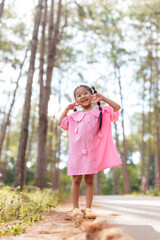 Asian girl in pink dress posing happily in forest. Smiling, showing peace signs with hands. Tall trees and natural light create bright, cheerful outdoor scene.
