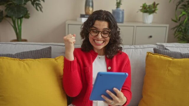 A young hispanic brunette woman smiling and celebrating with a clenched fist in her apartment living room, holding a tablet.