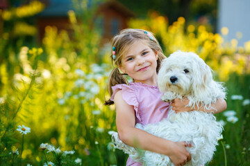 Adorable cute little girl and her puppy on the nature in the summer. Happy school child with eyeglasses holding Maltese dog, having fun with playing. Bright sunset light, active kid.