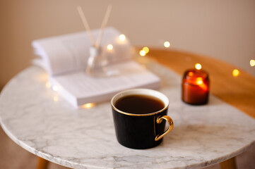 Cup of tea with open paper book and liquid home diffuser in glass bottle and bamboo sticks staying on marble coffee table close up. Cozy hygge atmosphere.