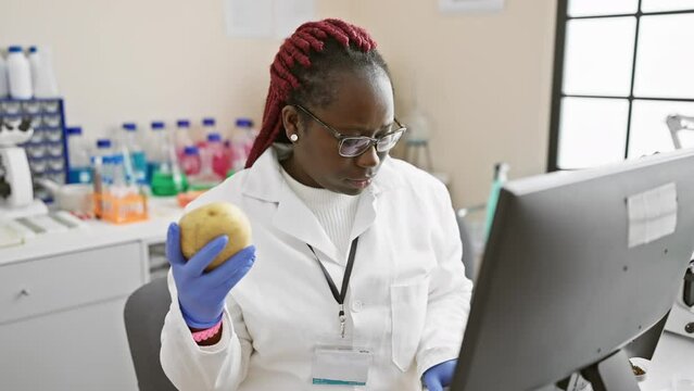 A focused woman with braids examining a potato in a laboratory, surrounded by scientific equipment.