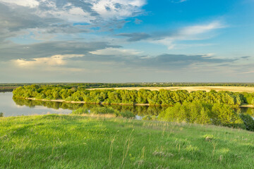 A scenic view of a river winding through a verdant landscape under a vibrant summer sky. The lush...