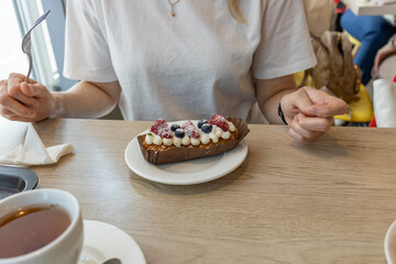 A person sits at a cafe table, enjoying a freshly baked cream puff topped with strawberries and blueberries. The table is set with a cup of tea and a napkin.