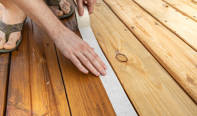 A close-up shot of a hand guiding painters tape across a wooden deck. The tape is being used to create a clean line for a painting project.