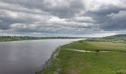 Fototapeta premium A wide river flows through a verdant landscape, bordered by lush green meadows and trees. The sky is a canvas of dramatic, gray clouds, creating a sense of grandeur and tranquility.