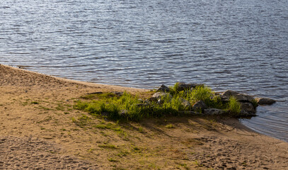 A sandy shoreline meets a calm lake at sunset, with a small patch of green grass and rocks breaking up the landscape. The water is a cool blue, reflecting the fading light of day.