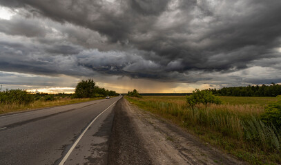 A lone road stretches towards the horizon, flanked by fields of green grass and a thick, dark line of storm clouds looming overhead.