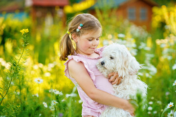 Adorable cute little girl and her puppy on the nature in the summer. Happy school child with eyeglasses holding Maltese dog, having fun with playing. Bright sunset light, active kid.
