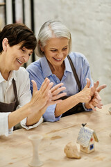 Two mature women, in cozy attire, are crafting together at an art studio, focusing on pottery.