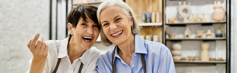 Two women, likely a couple, laugh together in a pottery studio, surrounded by their creations.