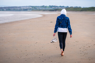 Sporty woman with sneakers in her hand on the sandy beach during windy day. Back view