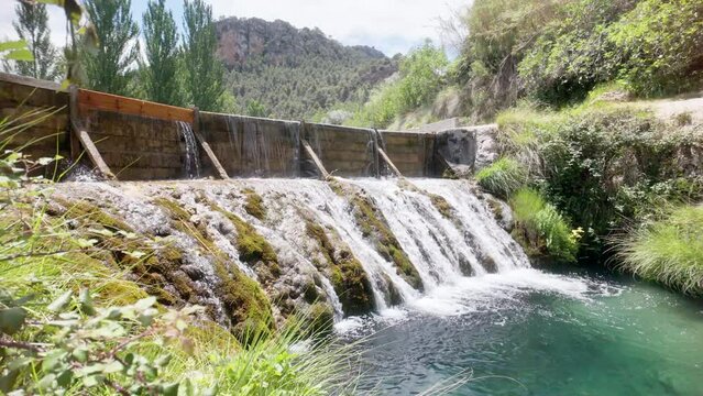 A waterfall is flowing into a river. The water is clear and the surrounding area is lush and green