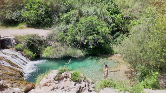 A woman is swimming in a clear pool of water. The water is surrounded by trees and rocks