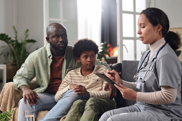 Side view portrait of young nurse consulting African American father and son during at home visit and holding clipboard copy space