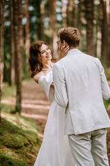 Fototapeta premium Valmiera, Latvia - September 9, 2023 - A smiling bride in a white dress touches the groom's face lovingly in a forest setting, with trees blurred in the background.