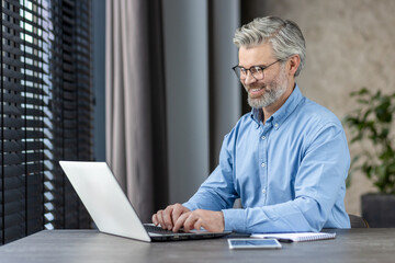 Fototapeta premium Smiling mature businessman working on laptop in a modern office setting. Concept of remote work, technology, and professional life.