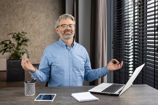 Confident mature man meditating at his office desk with a laptop and tablet, promoting mindfulness and relaxation in a work environment.