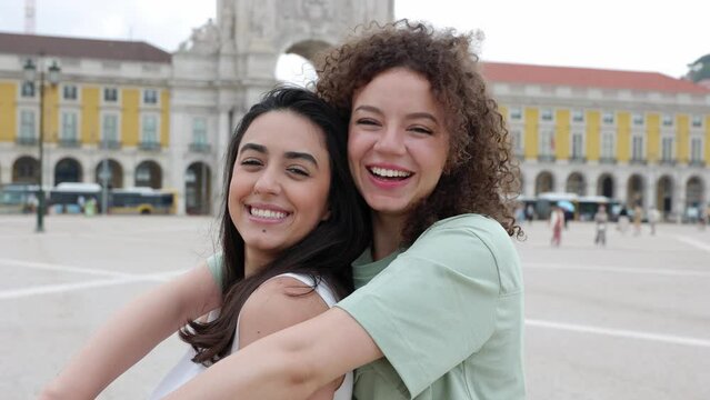 Portrait of two young female best friends smiling at camera standing at city street, Lisbon - Portugal. Happy millennial women hugging each other standing outdoors. Friendship and travel concept.