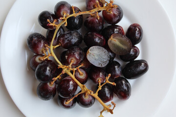 Dark purple or black grape with stem, on white plate, isolated on white background, flat lay or top view