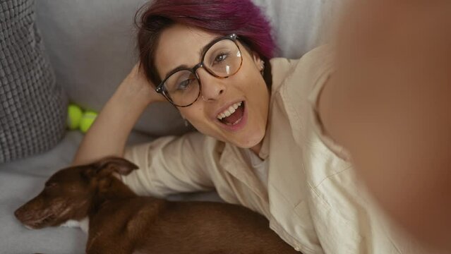 A young, beautiful woman with glasses and purple hair takes a fun selfie in her living room while her brown dog sleeps on the couch beside her.