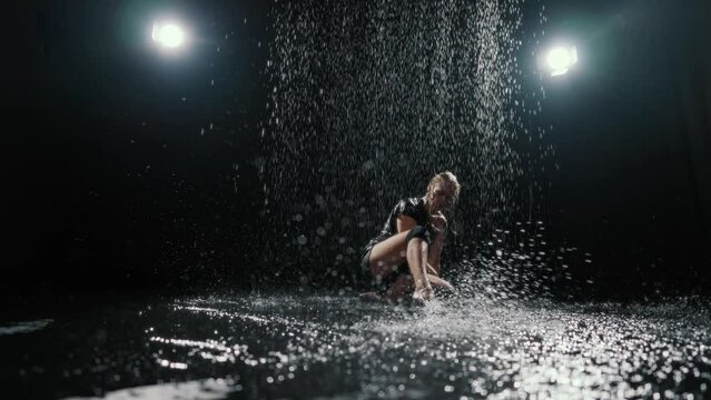 Passionate woman playing with water in dark studio, water flows in background. Graceful lady sitting on floor and splashing water, amazing slow motion shot, emotional female person in bright soffits