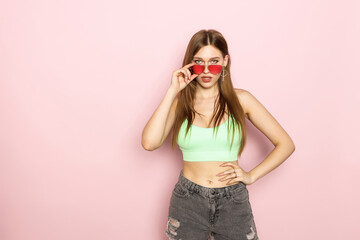 A woman is holding glasses and wearing a green top and shorts on a pink background. Beautiful figure and smile. Sexy look. Look from under glasses.